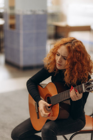 A woman with red curly hair plays a guitar with focus and expression in a comfortable environment. The scene evokes themes of music passion, creativity, and personal expression in an intimate setting.の写真素材