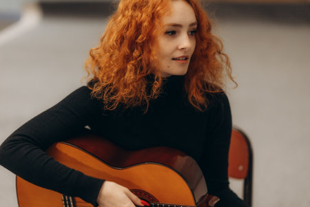 A young woman with curly red hair playing an acoustic guitar, wearing a black top, sitting indoors. She appears calm and focused, creating a cozy and artistic atmosphere.の写真素材