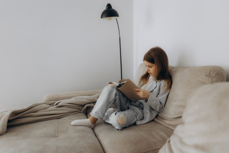 A young girl is seated comfortably on a beige sofa, engrossed in reading a book, enveloped by a minimalist and serene ambiance. She enjoys solitude amidst cozy surroundings highlighting a love for literature.の写真素材