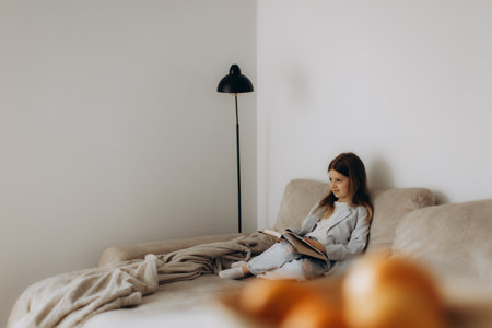 A young girl wearing casual clothes sits on a beige sofa reading a book. The room features a minimalist style, with soft lighting and decor creating a peaceful and cozy ambiance.の写真素材