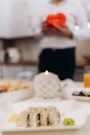 A close-up view of a sushi platter with a candlelit setting, depicting a romantic atmosphere. A blurred background shows someone holding a red gift, reinforcing the intimate mood.の写真素材