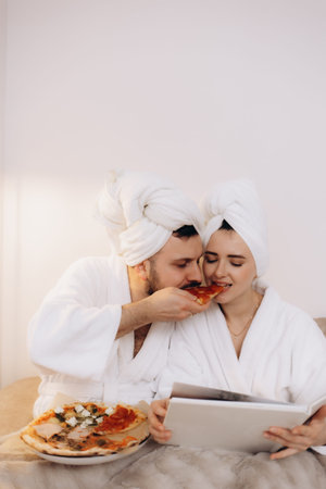 A smiling couple in matching bathrobes shares a pizza in a cozy setting, exuding love, leisure, and togetherness, emphasizing the joy of simple pleasures and quality time.の写真素材