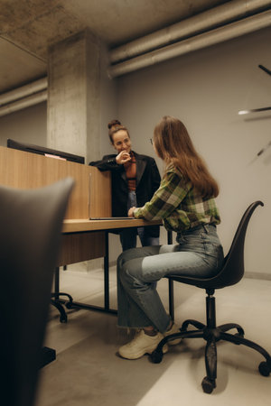 Two women are communicating in a contemporary workspace, sharing ideas while seated at a desk,...