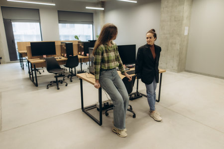 Two women interact casually by a desk in a contemporary office featuring modern workstations and an open design concept, fostering collaboration and professional connections within a serene and productive setting.の写真素材