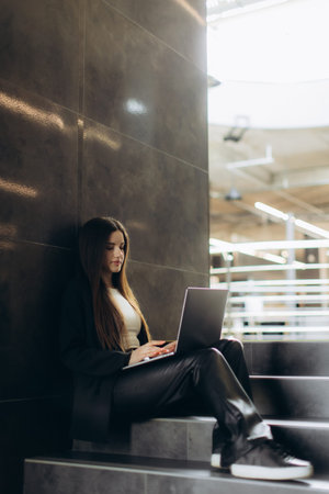 A young professional woman sits on steps in a modern office, working on her laptop. Sunlight filters through the windows, highlighting her focused expression and the contemporary architecture surrounding her.の写真素材