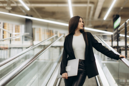 Professional woman in formal attire holding a laptop on an escalator. Bright modern lighting enhances the scene. Captures a corporate, poised vibe suitable for business and professionalism themes.の写真素材