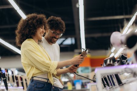 A cheerful couple selecting hair dryers in a modern electronics store, sharing a moment of joy while...