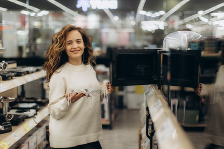 A young woman stands in an electronics store, smiling while selecting a microwave. Bright and modern shopping environment.の写真素材
