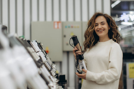 A cheerful young woman with curly hair choosing products in an electronics store, holding a blender and browsing a variety of gadgets. Her happy expression conveys excitement and satisfaction during her shopping experience.の写真素材