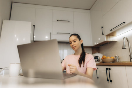 A young woman in a pink shirt works on a laptop at her kitchen table, surrounded by clean and modern...