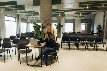 Two women working together on a laptop in a spacious, contemporary conference room setting...