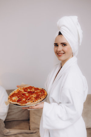 A woman wearing a white bathrobe and towel turban smiles while holding a fresh pepperoni pizza. Capturing a cozy and inviting atmosphere, she radiates comfort and relaxation in her home setting.の写真素材
