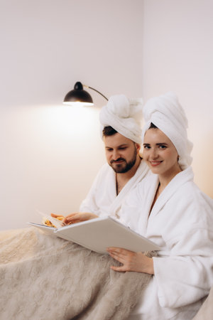 A cheerful couple in bathrobes relaxing, enjoying snacks, sharing a book, and savoring leisure time together under cozy lighting. The scene evokes calmness, comfort, warmth, and a sense of intimate companionship.の写真素材
