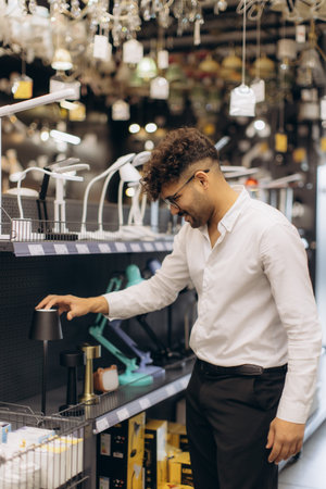 A man dressed in casual business attire examines a desk lamp in a lighting store, with shelves stocked with various lamp and decor options. The environment is well-lit with chandeliers overhead.の写真素材