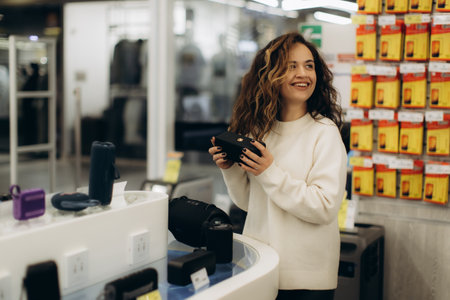 Smiling young woman browsing electronics in a retail store, considering various devices for purchase. The store is organized and vibrant, with displays showing product options and promotional offers.の写真素材