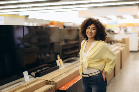 A cheerful woman standing near televisions in an electronics store, posing confidently and smiling warmly. Brightly lit environment with neatly arranged products emphasize a modern and professional retail atmosphere.の写真素材