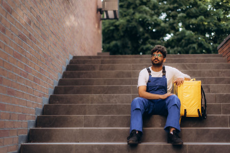 Tired delivery man resting on stairs, taking a break from delivering food with an insulated thermal backpack by his sideの写真素材