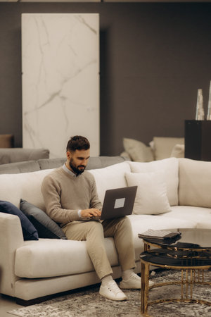 A person uses a laptop while seated on a cozy light-colored sofa within a modern living space,...