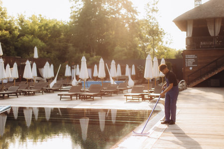 Maintenance worker cleaning a sparkling swimming pool at sunrise, surrounded by lush tropical scenery in a luxurious resort settingの写真素材