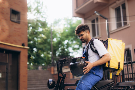 Courier wearing thermal backpack holding helmet while sitting on electric scooter, delivering food in urban city environmentの写真素材