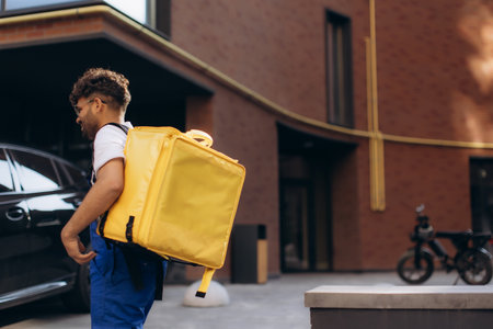 Courier carrying a yellow thermal backpack, delivering food in the city while walking near a parked car, showcasing urban logistics in actionの写真素材