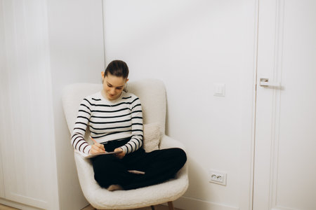A serene setting of a woman sitting in a cozy chair and writing in a notebook. She appears focused and relaxed, in a clean, minimalistic interior with neutral colors.の写真素材