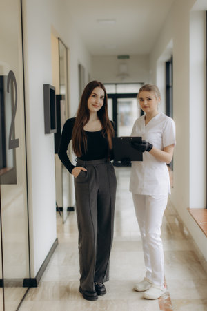 Two professional women standing in a bright office hallway, discussing documents and collaborating effectively while exuding professionalism and confidence, emphasizing a modern and organized work environment.の写真素材