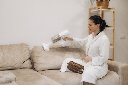 A woman wearing a bathrobe cleans her beige sofa using a handheld vacuum cleaner, emphasizing cleanliness and comfort in a cozy home environment.の写真素材