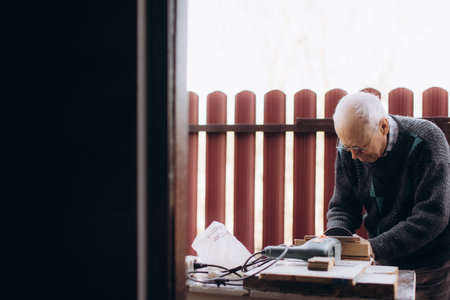 An elderly man focuses intently while working on a woodworking project outdoors, utilizing tools and materials. The background showcases a wooden fence and natural daylight, emphasizing craftsmanship and dedication.の写真素材