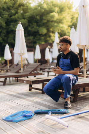 Pool cleaner using smartphone while sitting on deck chair near cleaning tools, taking a break from...