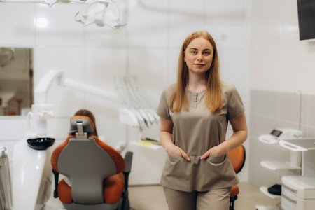 Female dentist standing in her modern dental clinic, with hands in pockets, while patient waits in dental chair in backgroundの写真素材