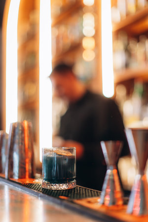 A sophisticated cocktail sits on a bartop, with blurred background details of bar equipment and lighting.の写真素材