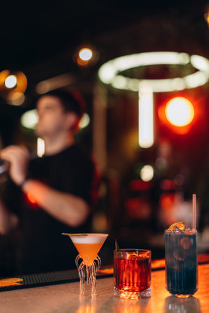 Three stylish cocktails are showcased on a bar counter, with blurred lighting creating a lively and sophisticated atmosphere. A bartender works in the background, enhancing the energetic and sociable vibe.の写真素材