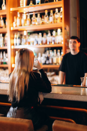 A woman sits at a stylish bar counter enjoying her drink, surrounded by bottles and ambiance.の写真素材