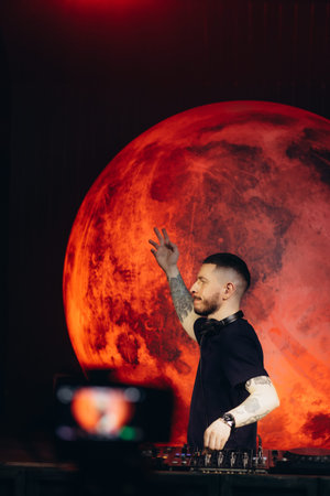 A DJ passionately performs on a set, mixing music while a bold crimson planet-like backdrop...