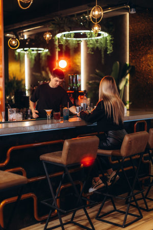 A warm evening scene in a modern bar as a bartender prepares drinks for a guest under atmospheric...