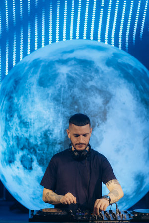 A professional DJ operates music equipment on stage, illuminated by a glowing large moon backdrop, under creative LED lighting, showcasing expertise and passion in music performance and live events.の写真素材
