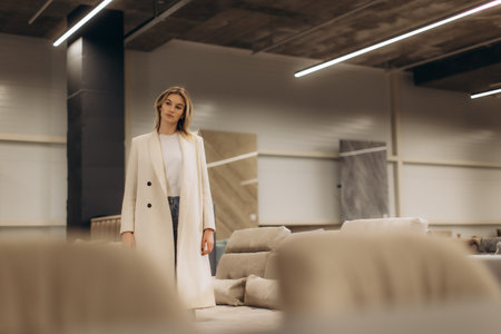 A fashionable young woman in a light coat stands confidently amidst contemporary furniture in a modern showroom, illustrating sophistication and style in interior environments.の写真素材