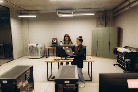 A workspace featuring two individuals cooperating on desktop and laptop computers. The contemporary office contains advanced equipment including printers and copiers, showcasing a collaborative work environment.の写真素材