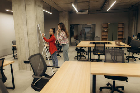 Two colleagues are working together on a whiteboard in an open, contemporary office space with desks, chairs, and a laptop in the background, fostering teamwork and creative collaboration in a professional environment.の写真素材