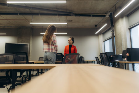 Two women are engaged in a conversation inside a modern office with minimalist decor. Bright lighting, sleek furniture, and open spaces define the professional environment.の写真素材