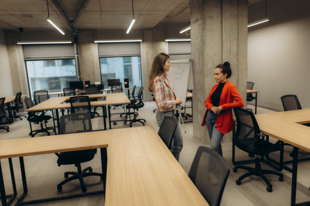 Two women are engaged in a discussion inside a spacious, modern office with wooden desks.の写真素材