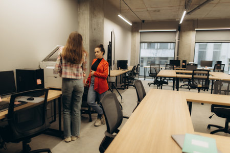 A modern office setup featuring two women by a printer, engaging in discussion, surrounded by workstations with computers on a sunny day.の写真素材