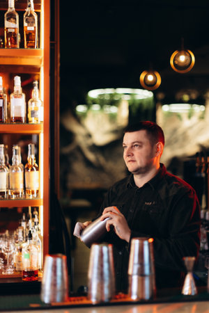 A bartender preparing drinks using a shaker in a modern bar setting with shelves of liquor bottles, illuminated by stylish warm lighting, creating an inviting nightlife ambiance.の写真素材