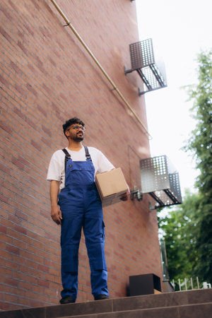 Delivery man carrying cardboard box and looking up while delivering package near brick buildingの写真素材