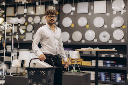 A man shopping for home lighting fixtures in a modern store. He is holding a shopping basket,...