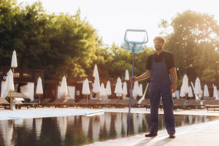 Pool cleaner holding cleaning net, standing by the poolside of a luxury hotel resort at sunset, ensuring clean and clear water for guestsの写真素材