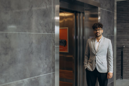 Professional young man standing by an elevator in a professional building, looking confident in a contemporary setting. Ideal for concepts like business, professionalism, urban lifestyle, or modern workspaces.の写真素材