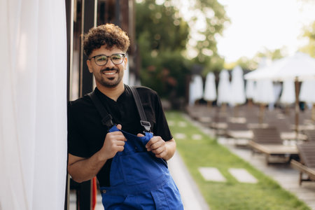 Portrait of a young handyman smiling and wearing blue overalls in a resort area with deck chairs and parasolsの写真素材