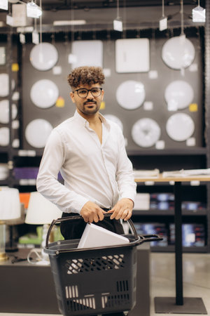 A man with glasses and a shirt holding a shopping basket stands in a lighting store surrounded by modern fixtures and lamps, showcasing an indoor shopping environment. Perfect for concepts like commerce and lifestyle.の写真素材